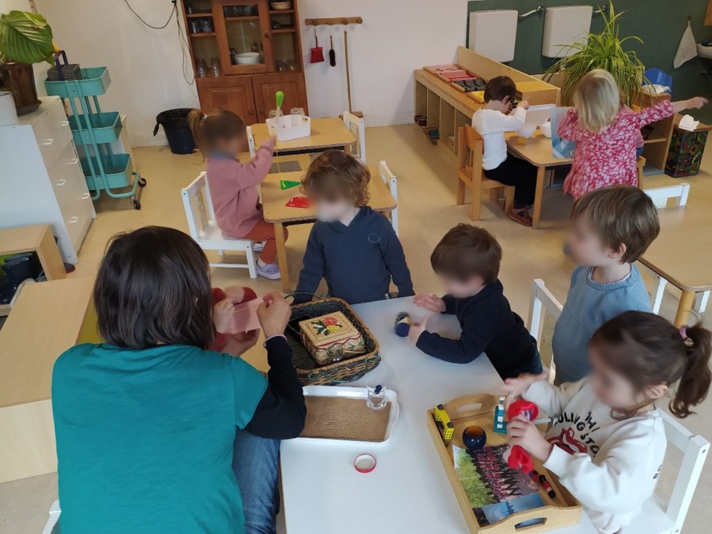Ensemble d'enfants qui composent la communauté enfantine du Village Montessori à Castelau-le-Lez. Ils sont assient un peu partout dans la classe mais un petit groupe s'est réuni autour de l'éducatrice pour regarder comment l'un des plus jeunes enfant utilise le matériel Montessori.