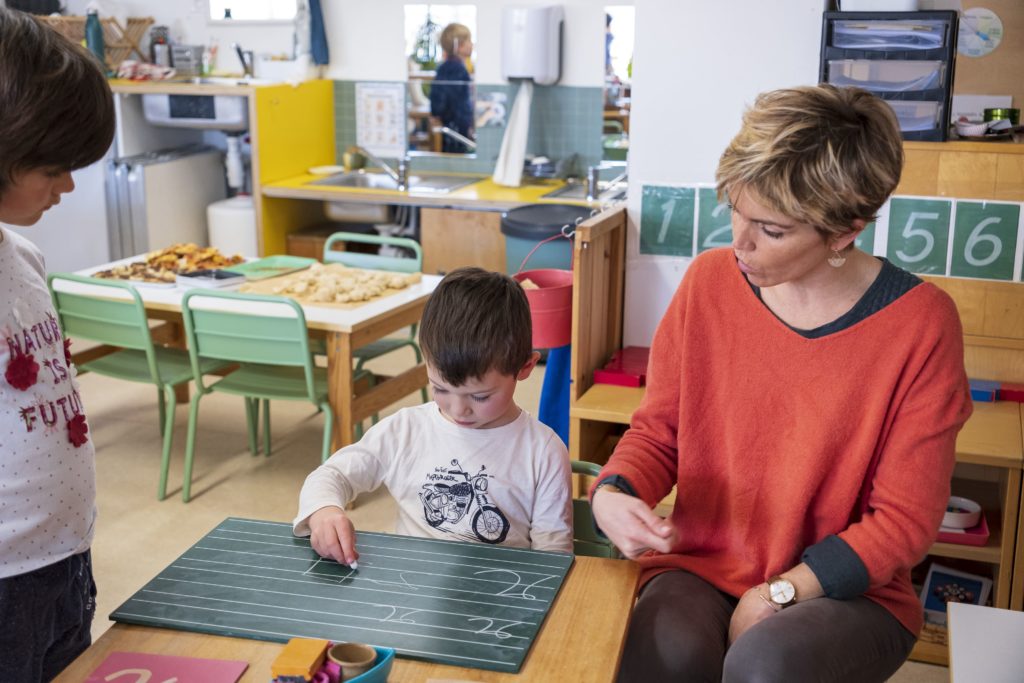 Une éducatrice de la pédagogie Montessori applique l'un des principe du décalogue de l'éducateur Montessori en montrant à l'enfant comment se servir du matériel Montessori. Ici il s'agit d'apprendre à écrire et donc avoir le bon geste pour exécuter cette tâche.
