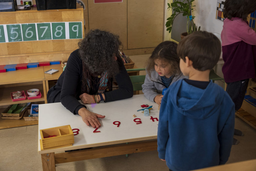 Une éducatrice Montessori 3/6 ans aide à la compréhension du matériel un jeune garçon. Comme dans le film documentaire, il apprend à reconnaitre les chiffres et l'éducatrice l'accompagne et le guide.